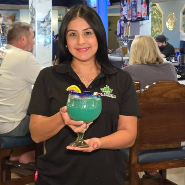 A smiling El Mezcal server holds a large blue margarita with a lemon slice in a lively restaurant.