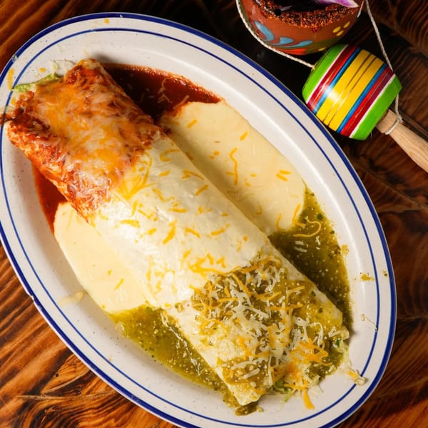 A large burrito covered in red sauce, queso, and green salsa, with a colorful maraca nearby.