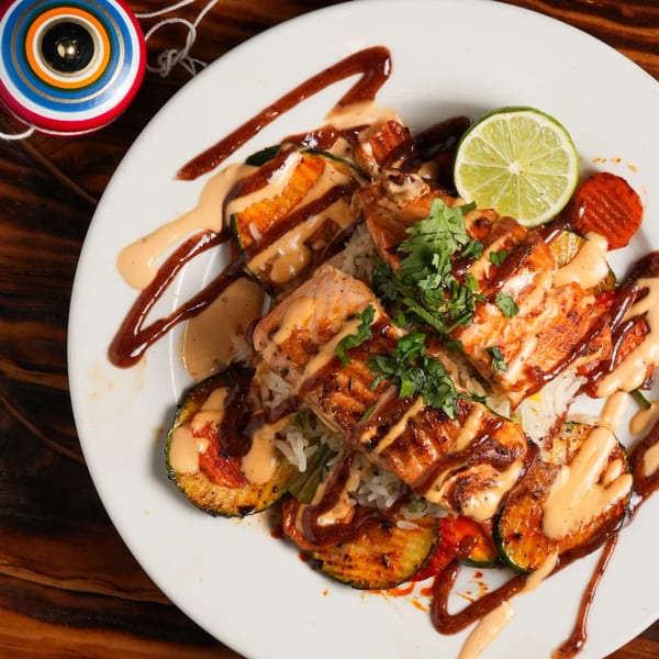 Grilled salmon with zucchini, carrots, rice, and sauces, garnished with cilantro and lime.