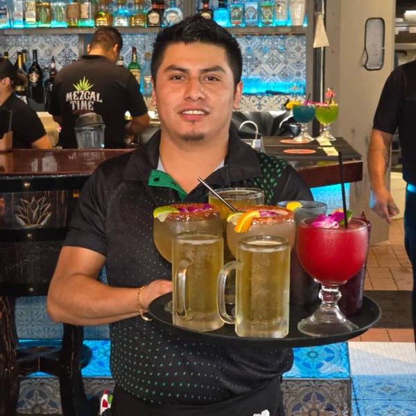 Smiling El Mezcal server holding a tray with four colorful drinks and two mugs of beer.