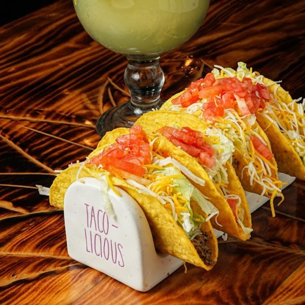 Four hard-shell tacos with ground beef, lettuce, cheese, and tomatoes, next to a margarita.