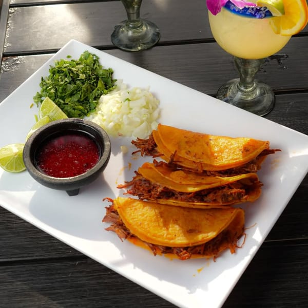 El Mezcal birria tacos with cilantro, onions, lime, and red sauce, next to a margarita.