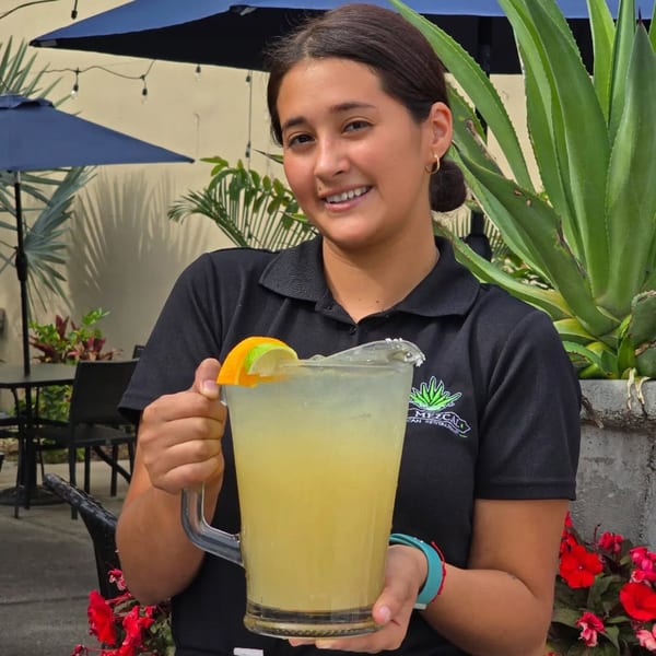 Smiling El Mezcal server holds a large pitcher of margarita on a sunny outdoor patio.