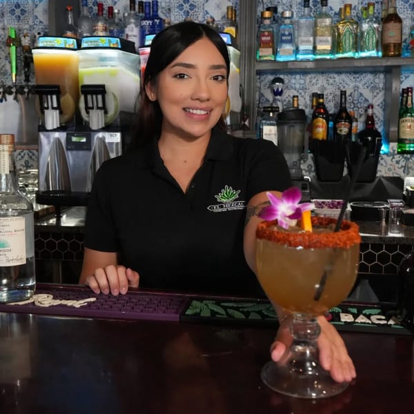 A smiling El Mezcal bartender presents a large margarita with a chili-lime rim and orchid garnish.