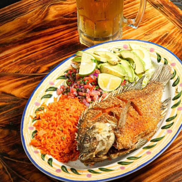 Fried whole fish with rice, pico de gallo, avocado, lime, and a beer on a wooden table.